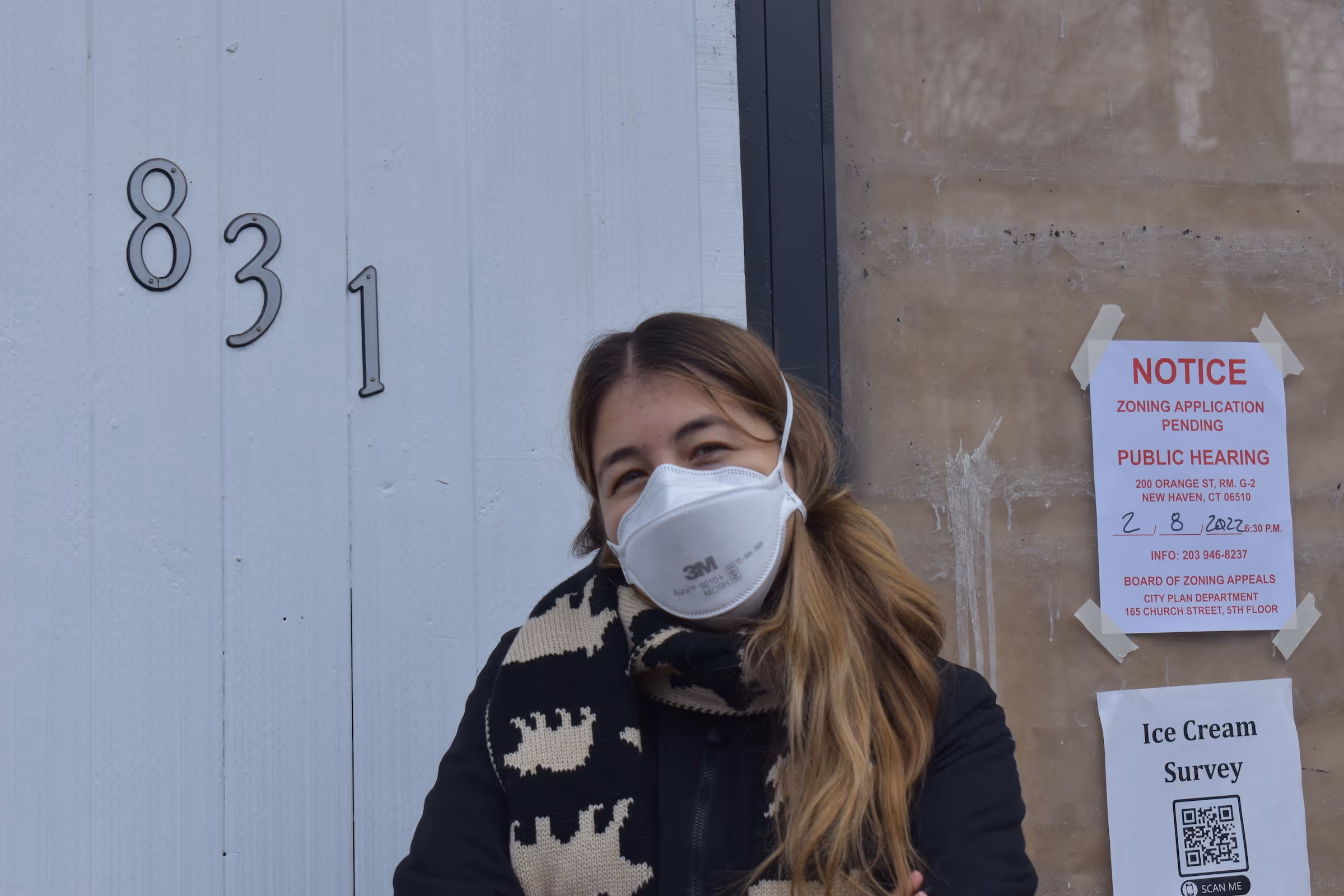 Elena outside the storefront: Elena wearing a mask and winter coat standing in front of her shop during the early stages of development. A zoning application notice is posted on the door along with an "Ice Cream Survey" sign with a QR code. Credit noted as from the New Haven Independent.