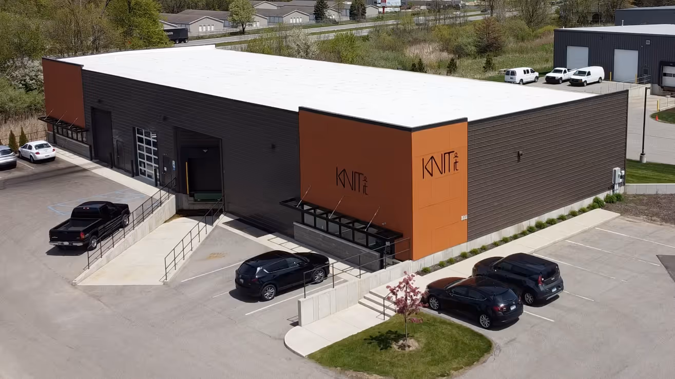 Aerial view of KnitIt's modern manufacturing facility in Holland, Michigan, showing a large industrial building with orange and dark metal siding, the KnitIt logo prominently displayed on the exterior, and a parking lot with several vehicles, demonstrating the company's commitment to local manufacturing.