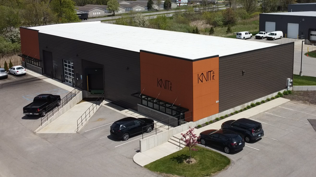 Aerial view of KnitIt's modern manufacturing facility in Holland, Michigan, showing a large industrial building with orange and dark metal siding, the KnitIt logo prominently displayed on the exterior, and a parking lot with several vehicles, demonstrating the company's commitment to local manufacturing.