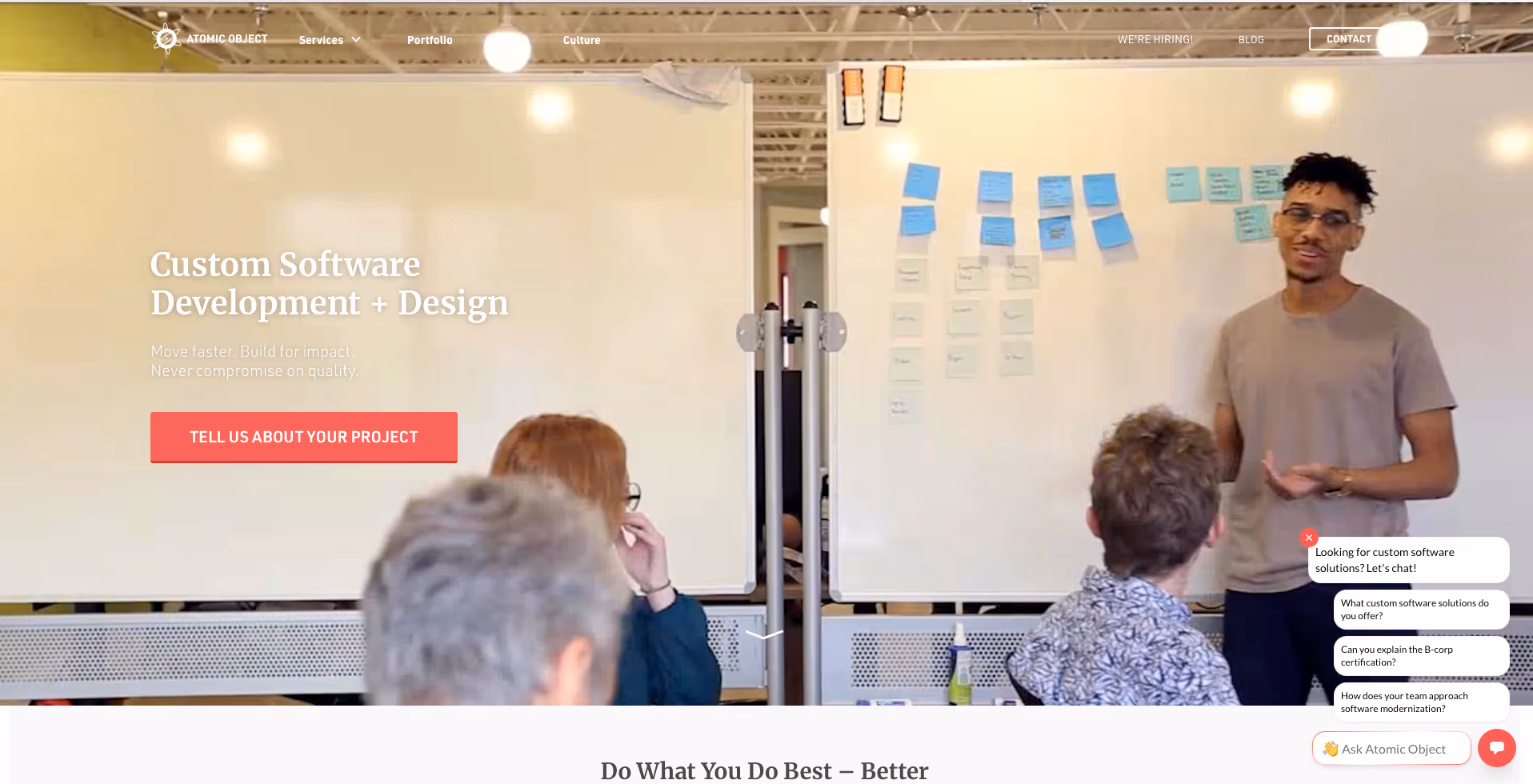 Atomic Object website homepage showing their office space with team members collaborating. A person in a tan shirt stands presenting to colleagues seated at a table, with blue sticky notes on the wall behind them. The page header reads "Custom Software Development + Design" with taglines "Move faster. Build for impact. Never compromise on quality." A red "Tell us about your project" button is visible, along with a chat widget in the bottom right corner.