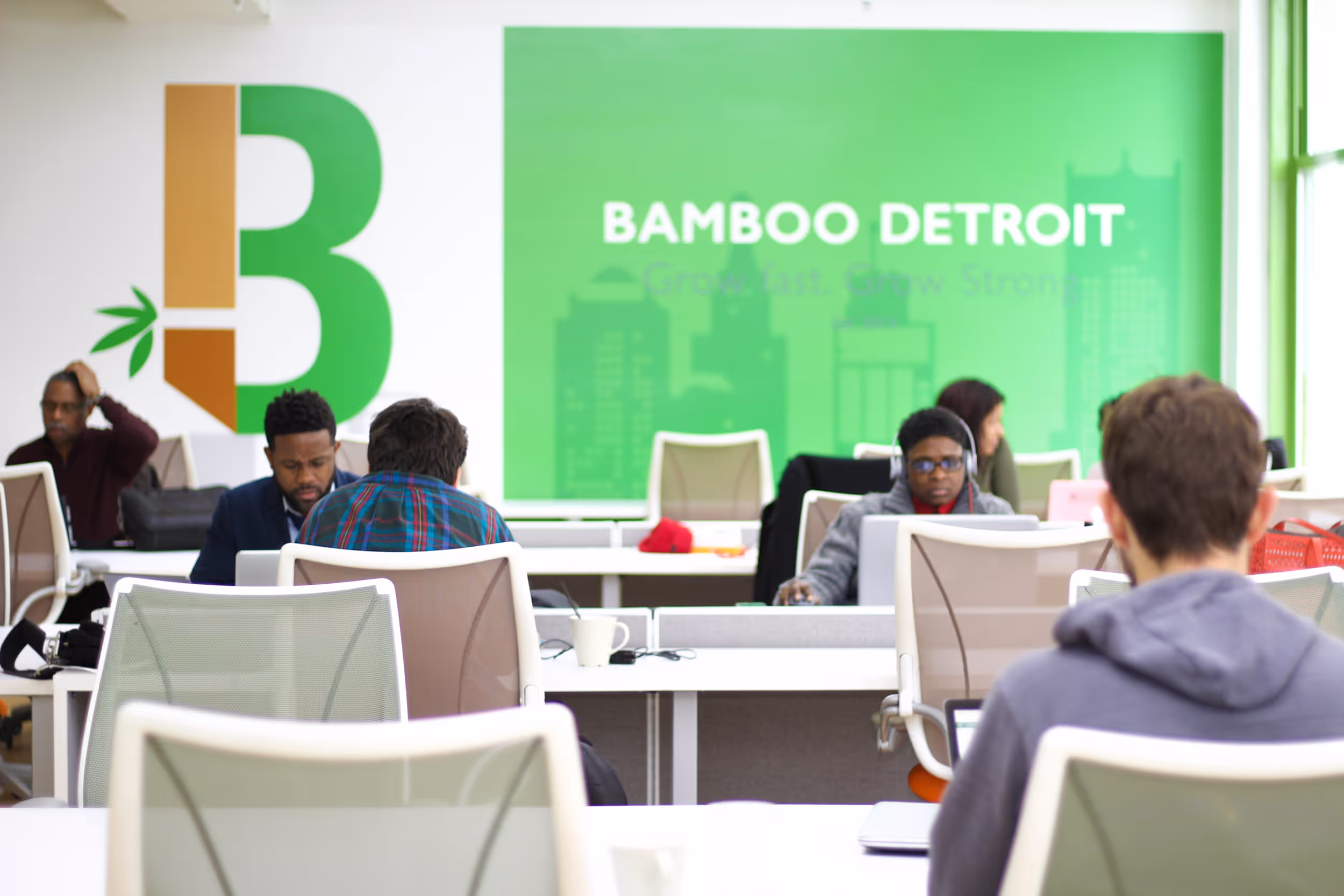 Interior of Bamboo Detroit co-working space showing an open office environment with white desks and chairs. Multiple people are working at laptops in the modern space, with a large green wall displaying 'BAMBOO DETROIT' branding and natural light coming through windows.