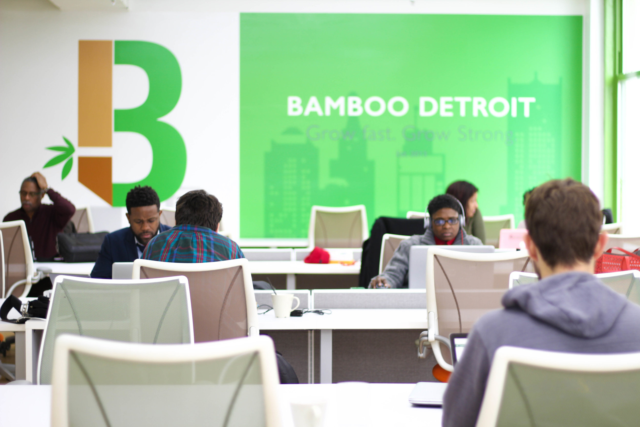 Interior of Bamboo Detroit co-working space showing an open office environment with white desks and chairs. Multiple people are working at laptops in the modern space, with a large green wall displaying 'BAMBOO DETROIT' branding and natural light coming through windows.