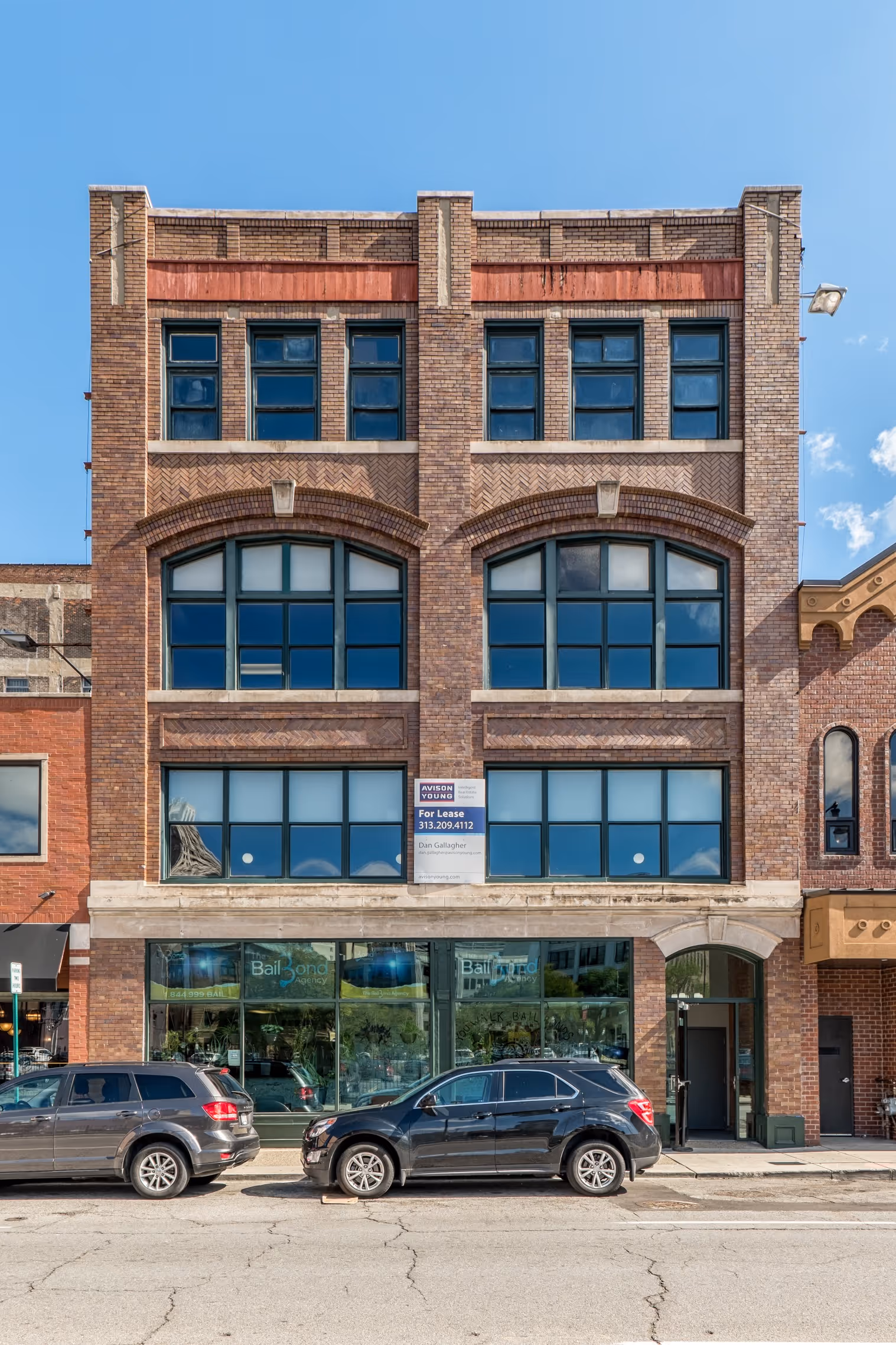 Four-story historic brick building in downtown Detroit with large arched windows on the second floor and rectangular windows above. The ground floor has retail storefronts, and there are cars parked on the street in front. A white leasing sign is visible in one of the upper floor windows.