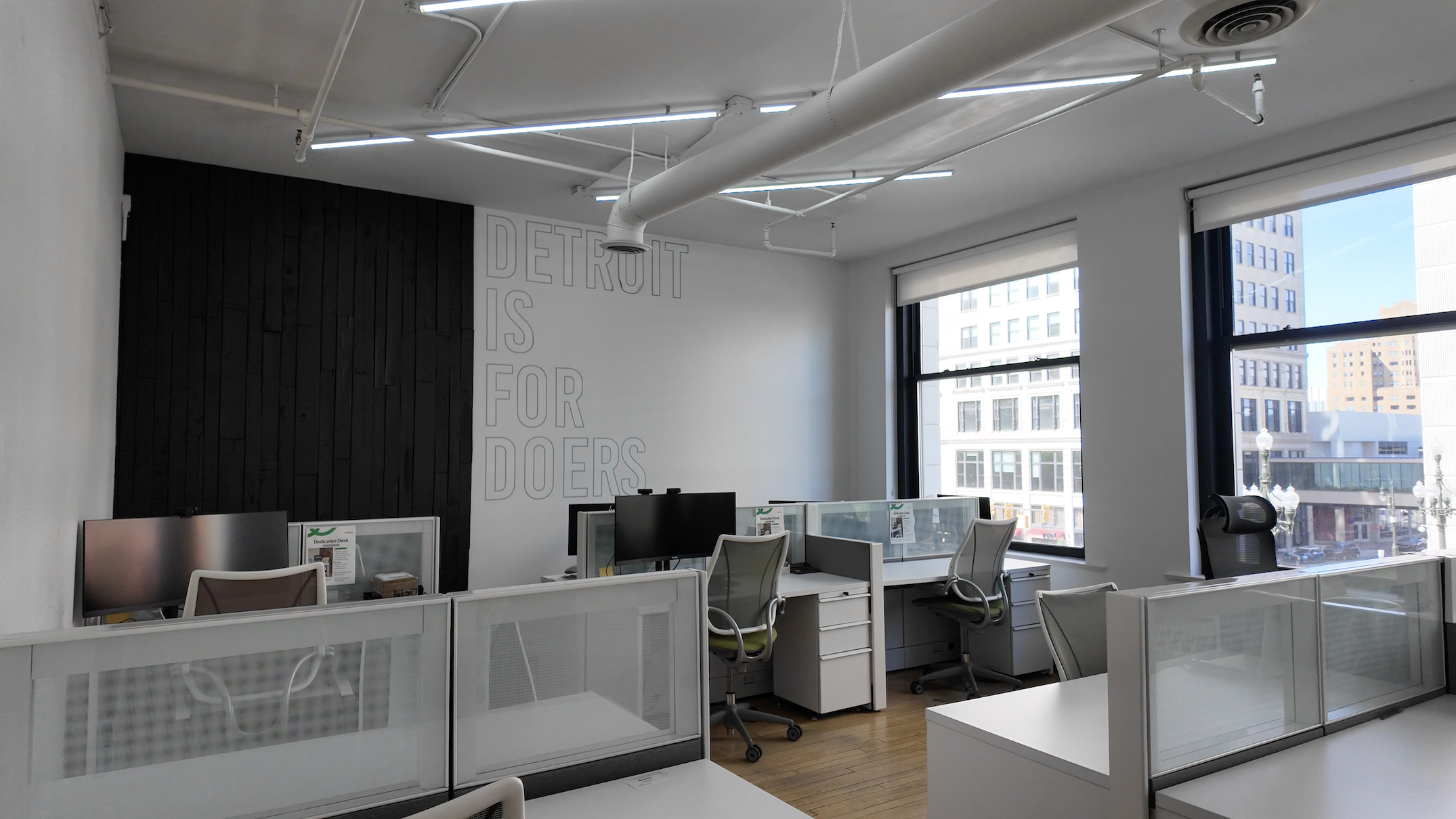 Modern office interior with an open floor plan featuring white workstations separated by glass partitions. The space has polished concrete floors, exposed ceiling with track lighting, and large windows. Text on the wall reads 'DETROIT IS FOR DOERS' in gray letters.