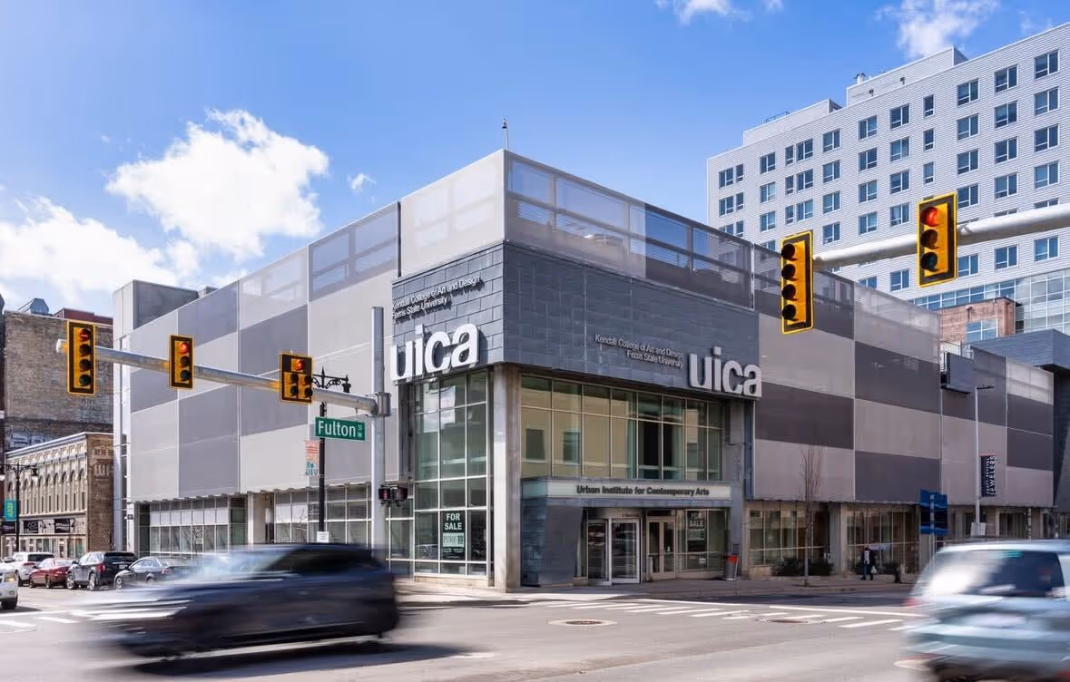 Large modern building with gray and white geometric exterior panels. The building has 'UICA' signage and sits at a busy intersection with traffic lights visible. Multiple cars are driving past the building, creating motion blur in the foreground.