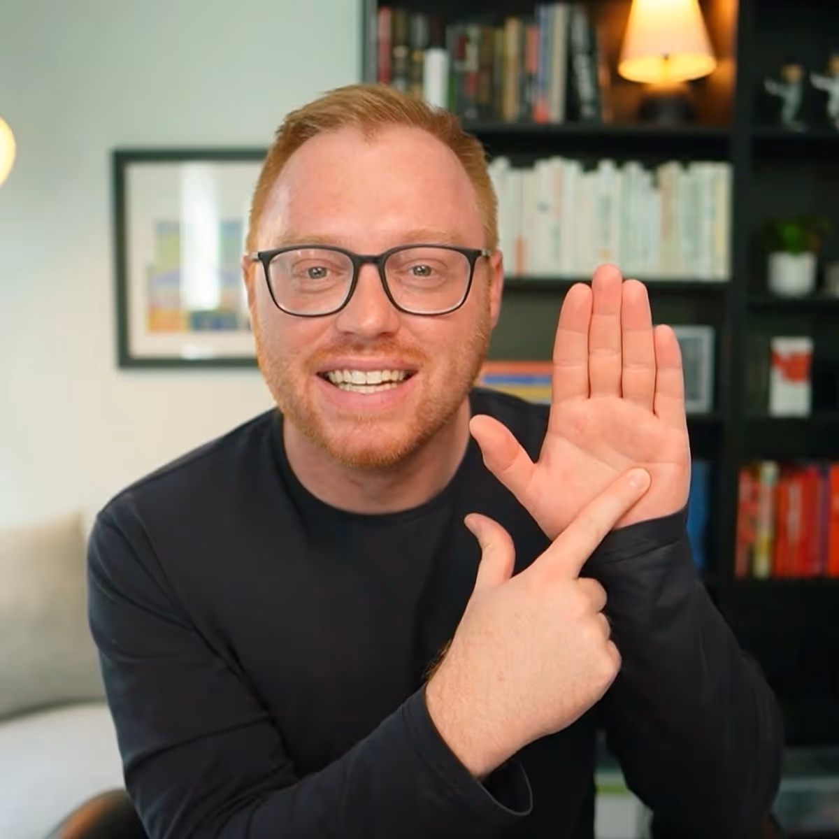 Man with glasses and reddish hair wearing a dark shirt, sitting and making a hand gesture with one hand raised and the other pointing. He's in what appears to be a home office or living room with bookshelves visible in the background.