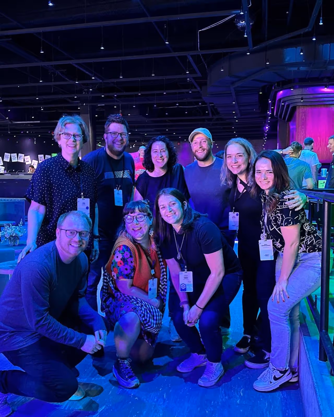 Group photo of nine people at a conference venue with purple stage lighting, wearing conference badges and casual attire, standing and kneeling together for a team photo.