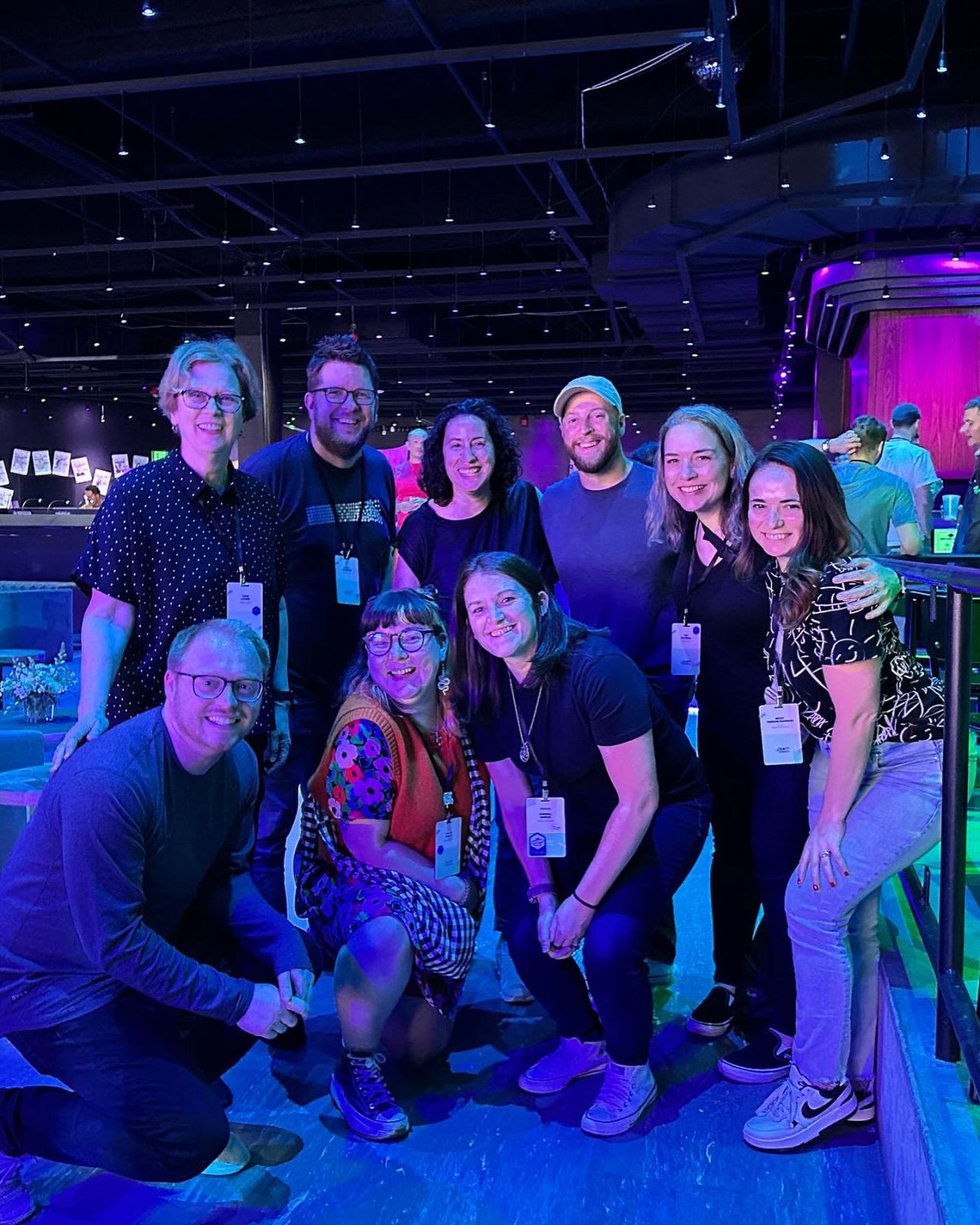 Group photo of nine people at a conference venue with purple stage lighting, wearing conference badges and casual attire, standing and kneeling together for a team photo.