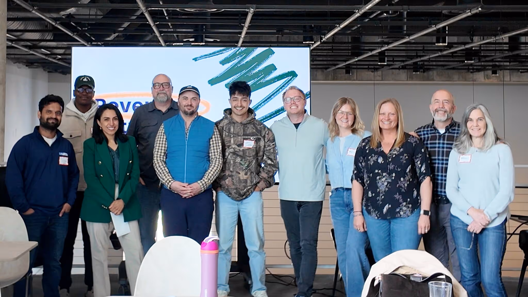 A group of eleven founders and entrepreneurs posing together in front of a large display screen showing "Revenue Retreat" branding at Bamboo Grand Rapids. The group stands in a modern coworking space with exposed ceiling infrastructure, round tables, and white chairs visible in the foreground. Most attendees are wearing name tags. The mood is warm and celebratory, suggesting an end-of-day group photo from The Quarter's May 2026 Revenue Retreat.