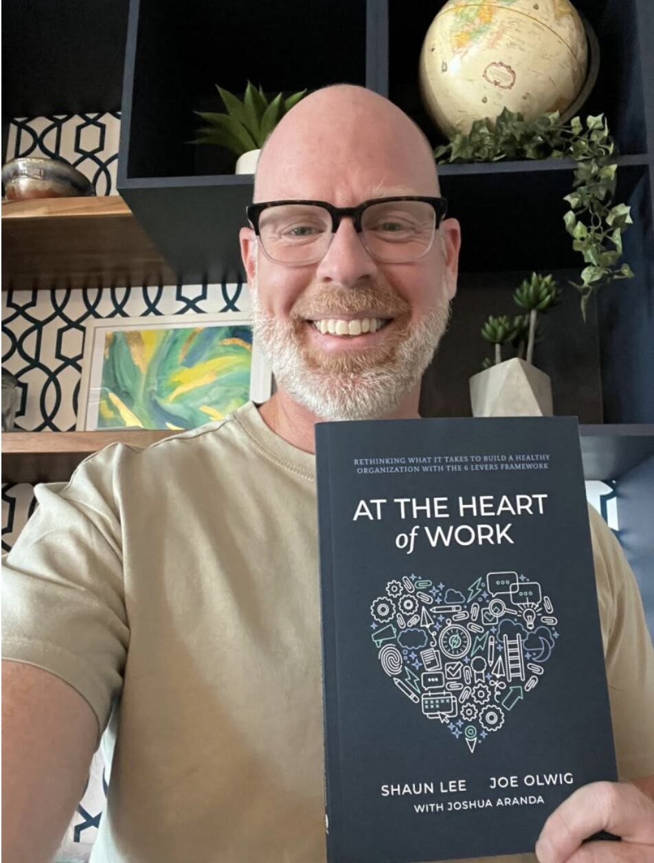 Shaun Lee holding a copy of his book At the Heart of Work, co-authored with Joe Olwig and Joshua Aranda, with a dark navy cover featuring a heart made of illustrated workplace icons.