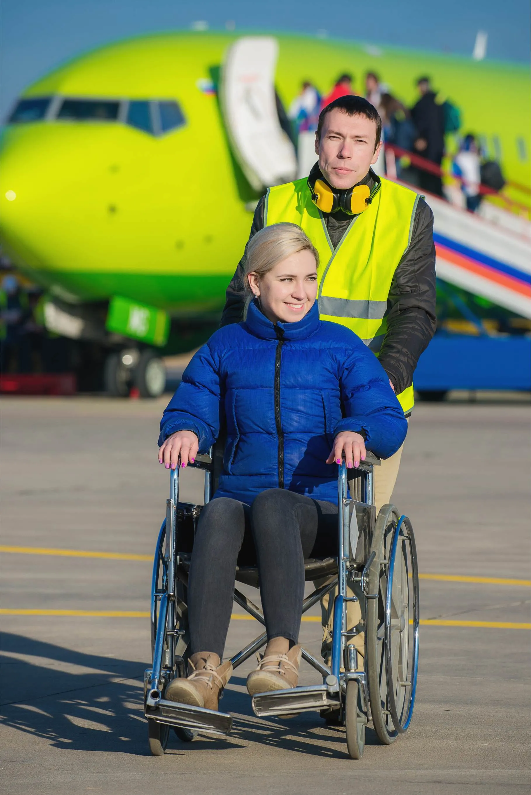 Woman in a wheelchair with airport assistant pushing the wheelchair away from a yellow aircraft.