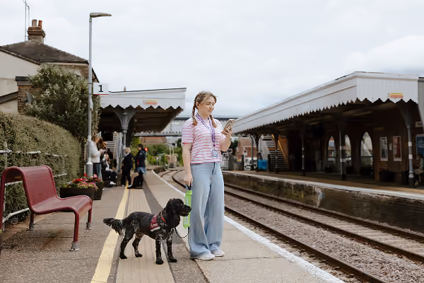 Photo of a lady with an assistance dog wearing a National Disability Card on a Verified Hidden Disability lanyard. She is looking at her phone and facing the railway.