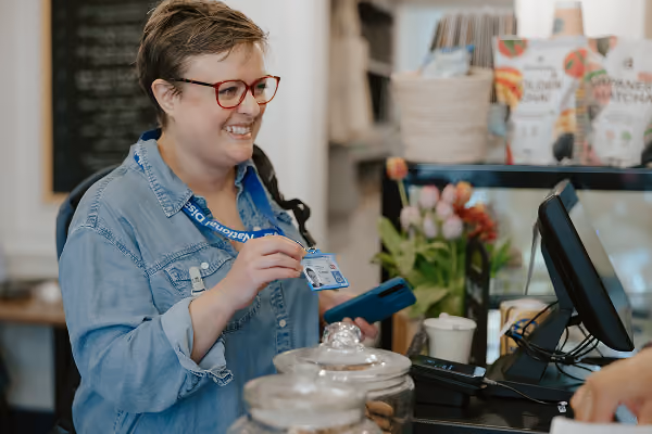 Photo of a woman in a denim shirt smiling and holding up her National Disability Card at a shop till