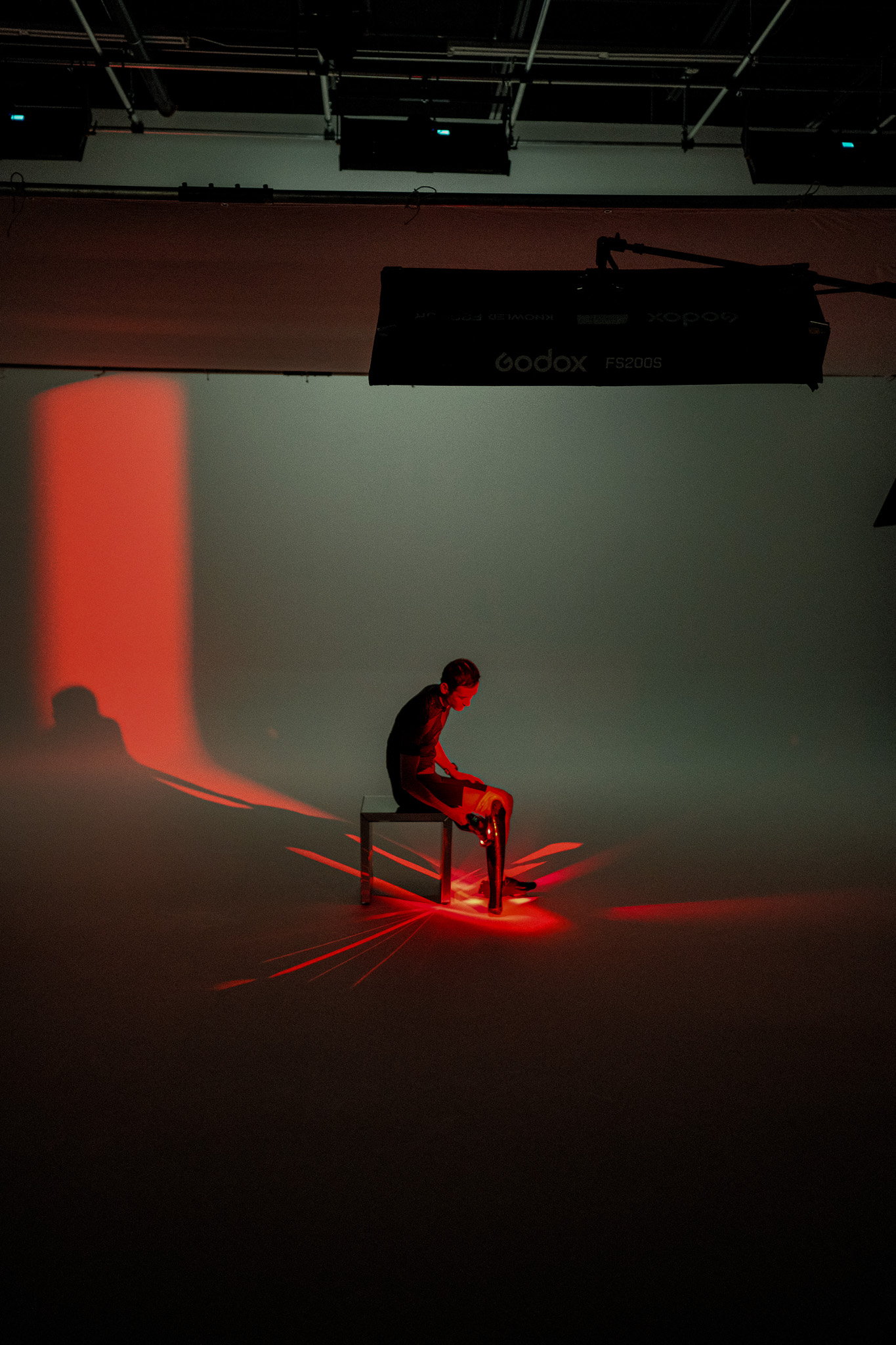 Kai seated alone in a shaft of red light using a Theragun on his leg