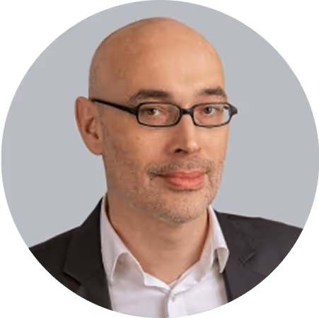 Ein Mann mit Brille und Glatze in einem Anzug steht vor einem grünen Hintergrund.