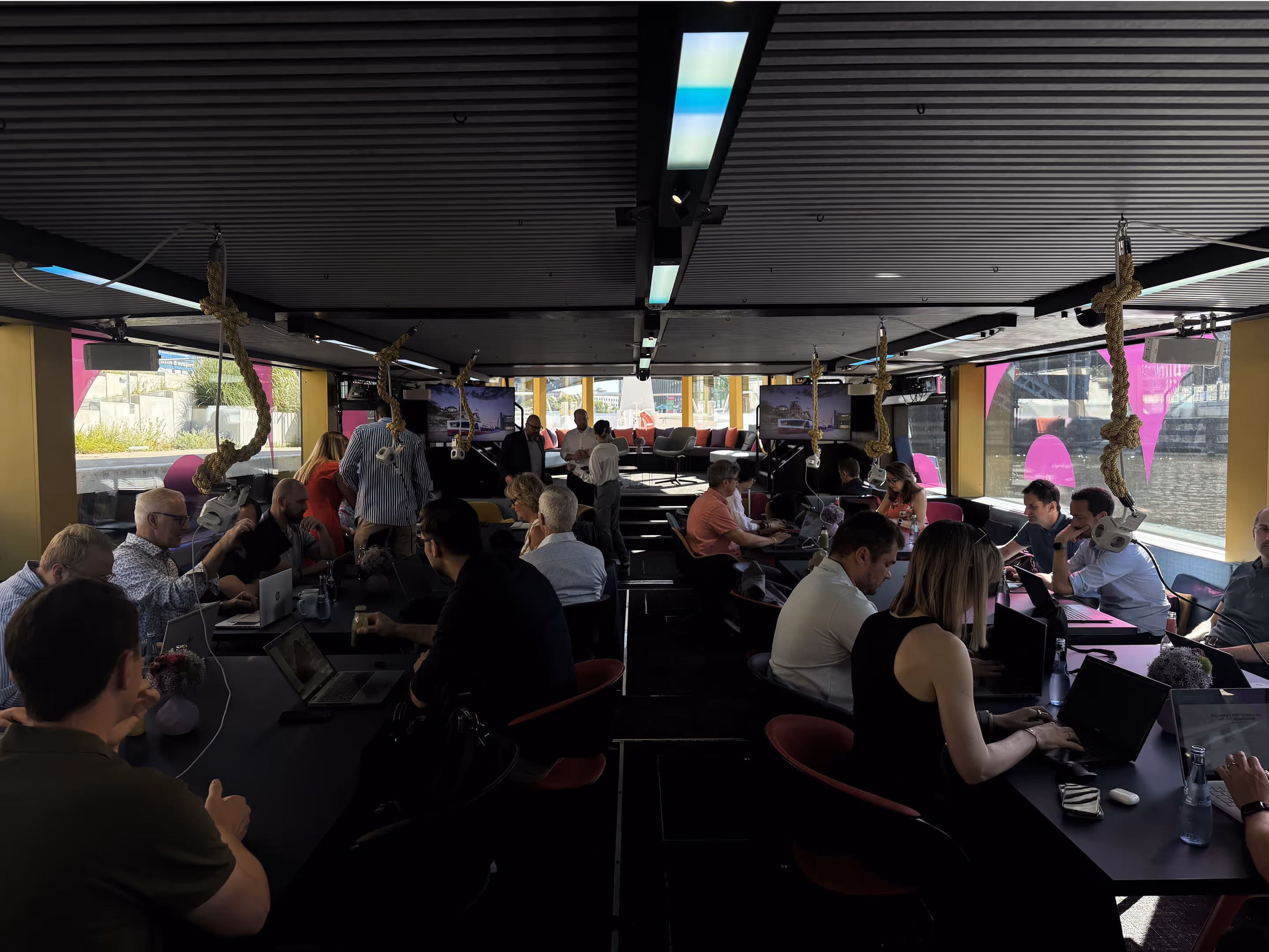 Mehrere Personen arbeiten an Computern in einem modernen Büro mit rosa Flaggen und Seilen im Hintergrund.