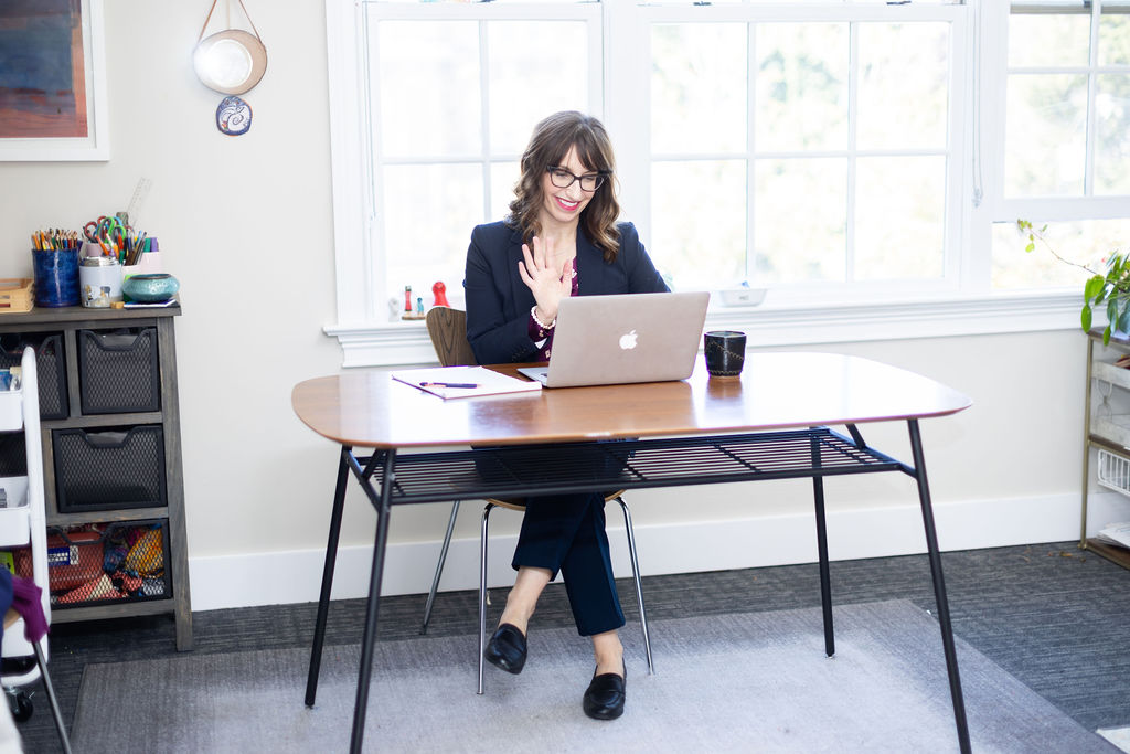 Dr. Jen sitting in a modern office waving to her computer screen for her virtual process group meeting.