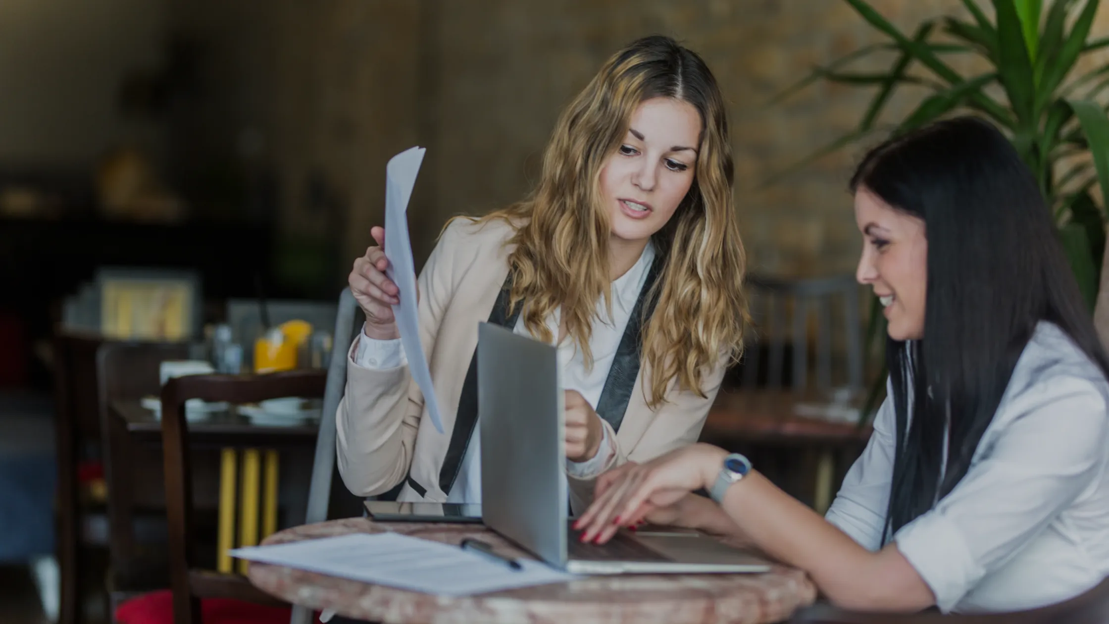 Two women are working together at a table, with one holding papers and the other pointing at a laptop screen, engaged in a discussion.