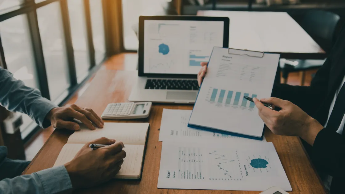 A business meeting in progress with individuals reviewing financial documents and a laptop displaying graphs.