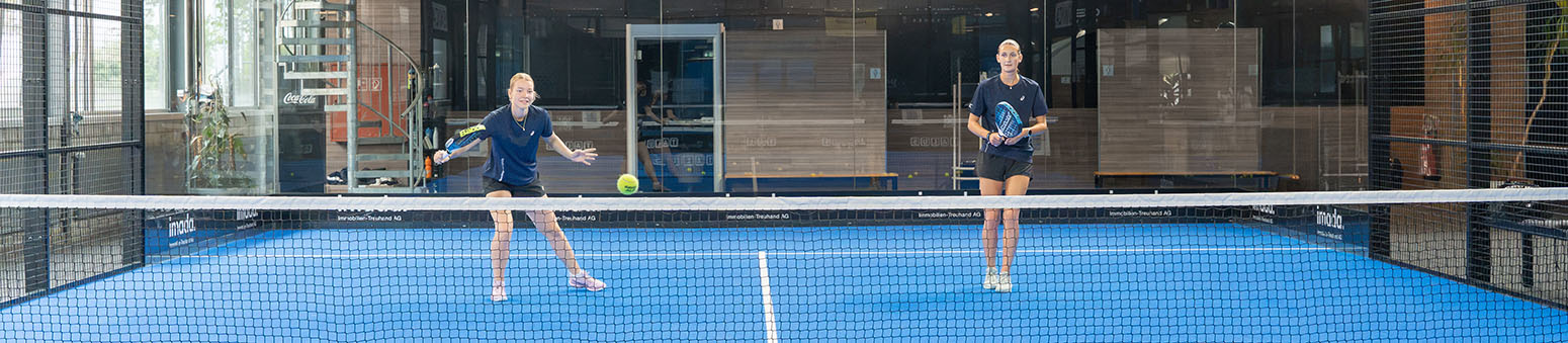 Zwei Frauen spielen Padel-Tennis auf einem blauem Innenplatz, eine schlägt einen Ball mit dem Schläger, die andere steht bereit.