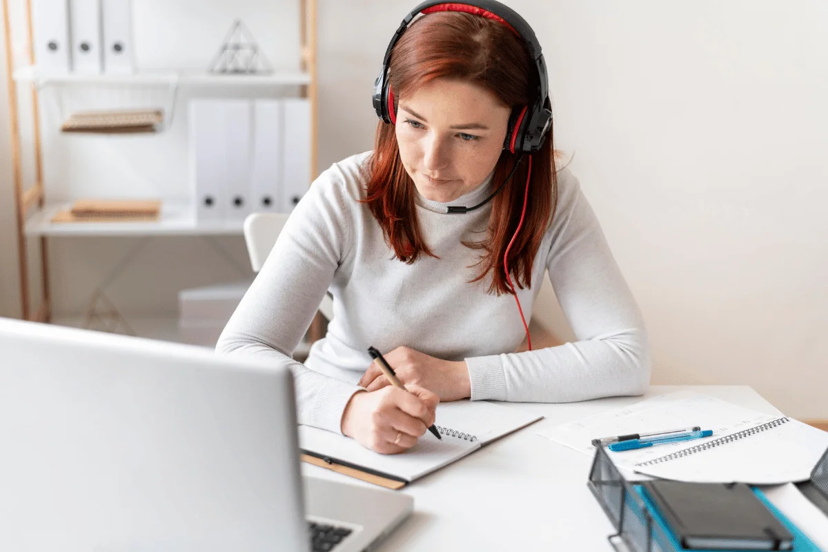 A woman with red hair wearing a headset is focused on writing in a notebook while watching a laptop. She appears engaged in a remote learning setting.