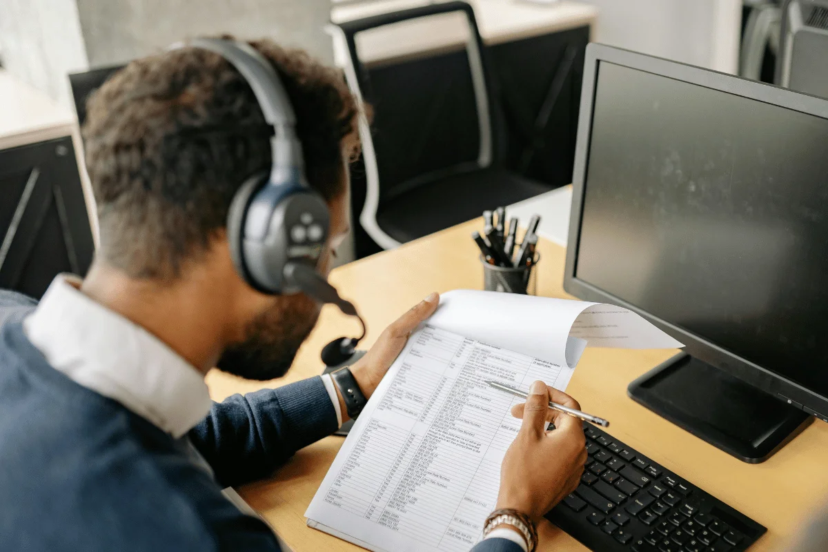A person wearing headphones sits at a desk, focused on a document filled with text. A keyboard and monitor are in front, conveying a work setting.