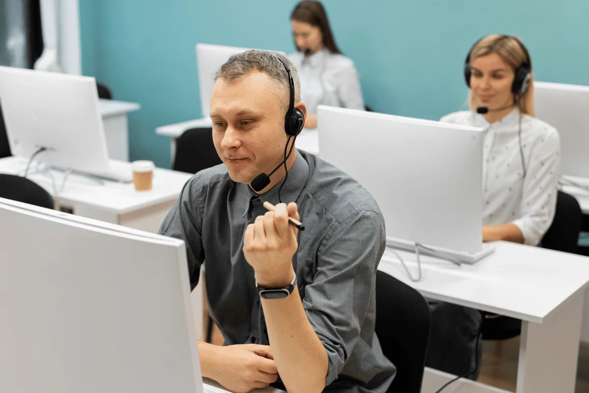 A man with a headset works at a computer in a modern call center with several colleagues. The atmosphere is focused and professional.