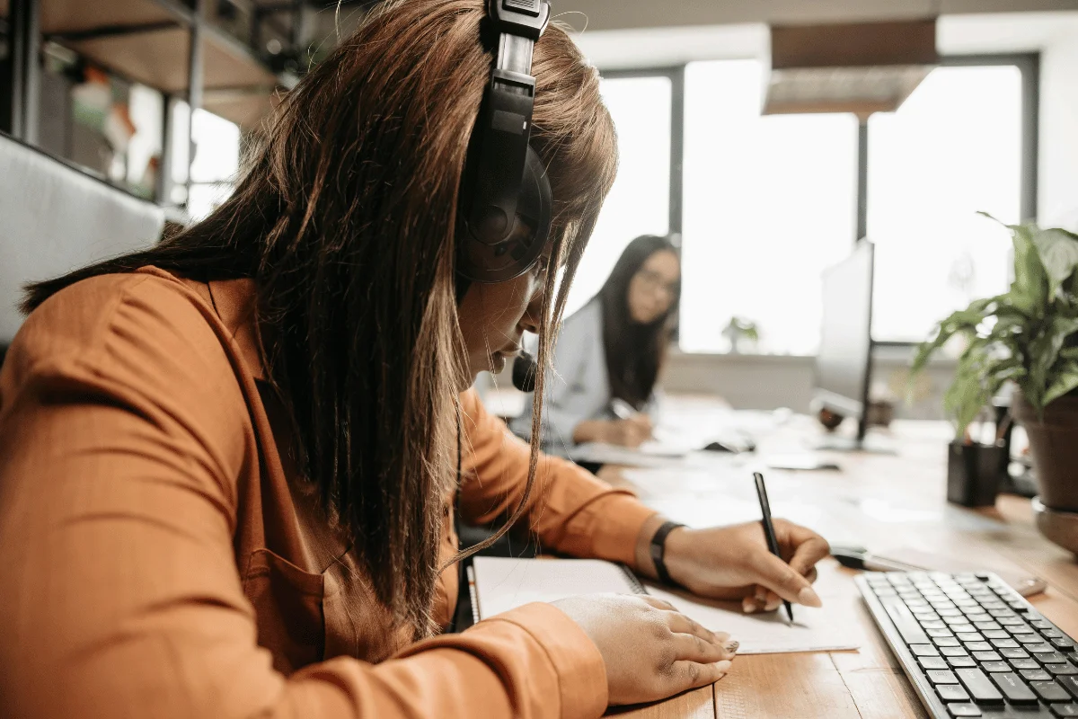 Female call center agent wearing headset writing notes at desk.