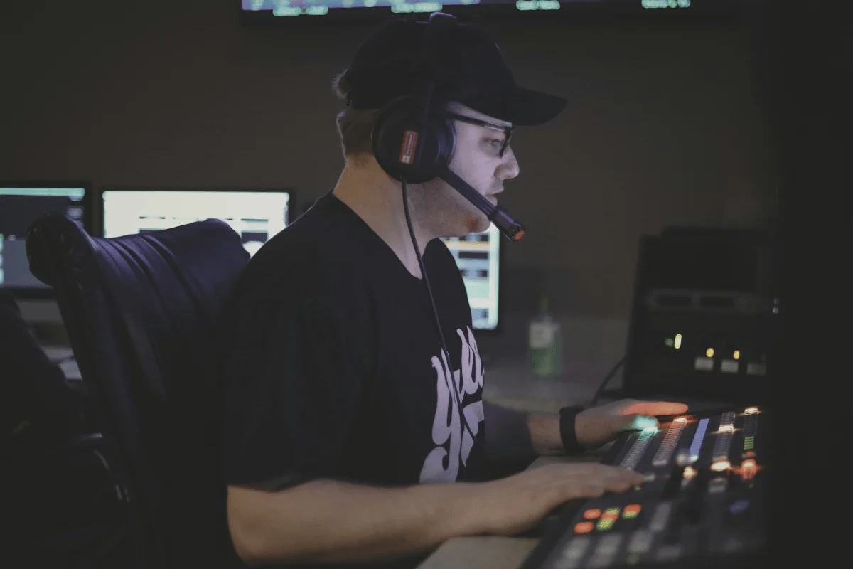 A person in a dark room wearing a headset operates a soundboard, focused on screens and controls, suggesting concentration in a broadcasting studio.