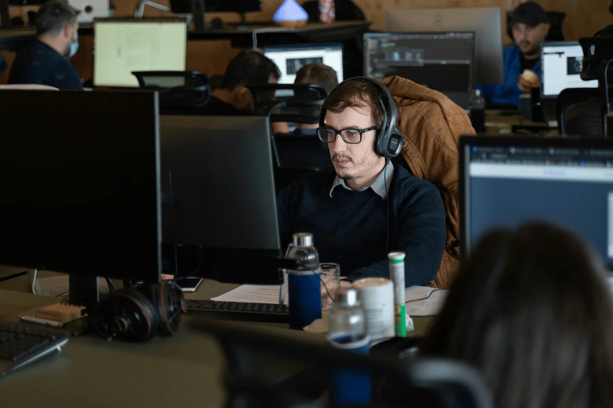 A man wearing headphones and glasses focuses intently on his computer screen in a busy office setting. The environment is collaborative and tech-oriented.
