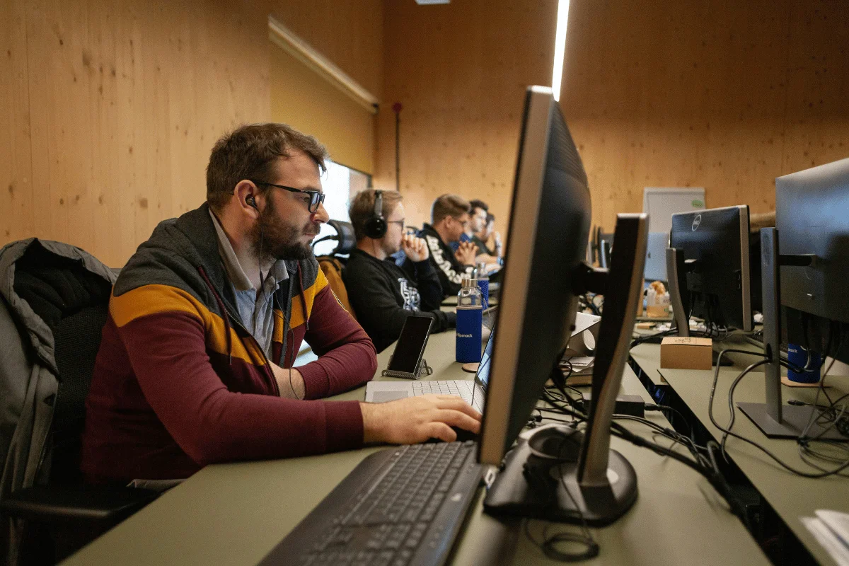 A group of focused individuals at a row of computers in an office setting. They wear headsets, implying a collaborative or tech-driven environment.