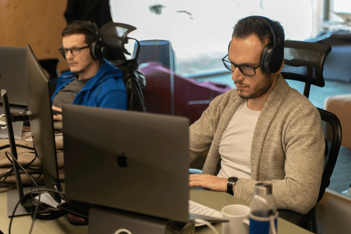 Two men focused on working at computers in an office, wearing headphones. The scene conveys a productive and concentrated atmosphere.