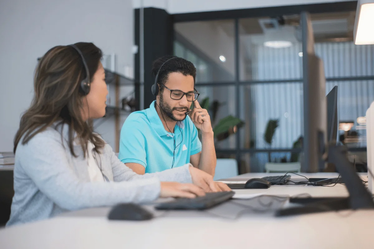 Two people wearing headsets sit in a modern office, focused on computer screens. The setting is collaborative, conveying a professional atmosphere.