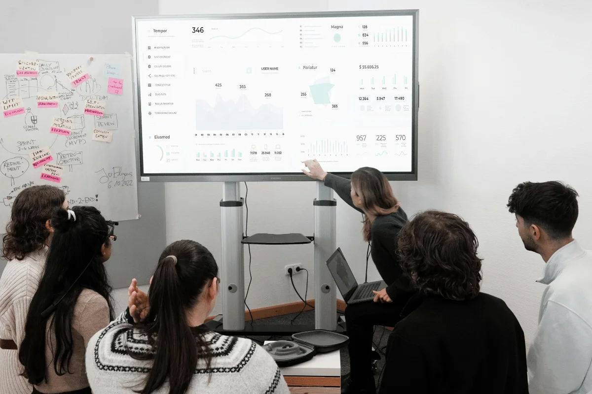 A group of six people sit around a screen showing data charts and graphs. A person points at the screen, explaining the data. A whiteboard with notes and sticky notes is in the background.