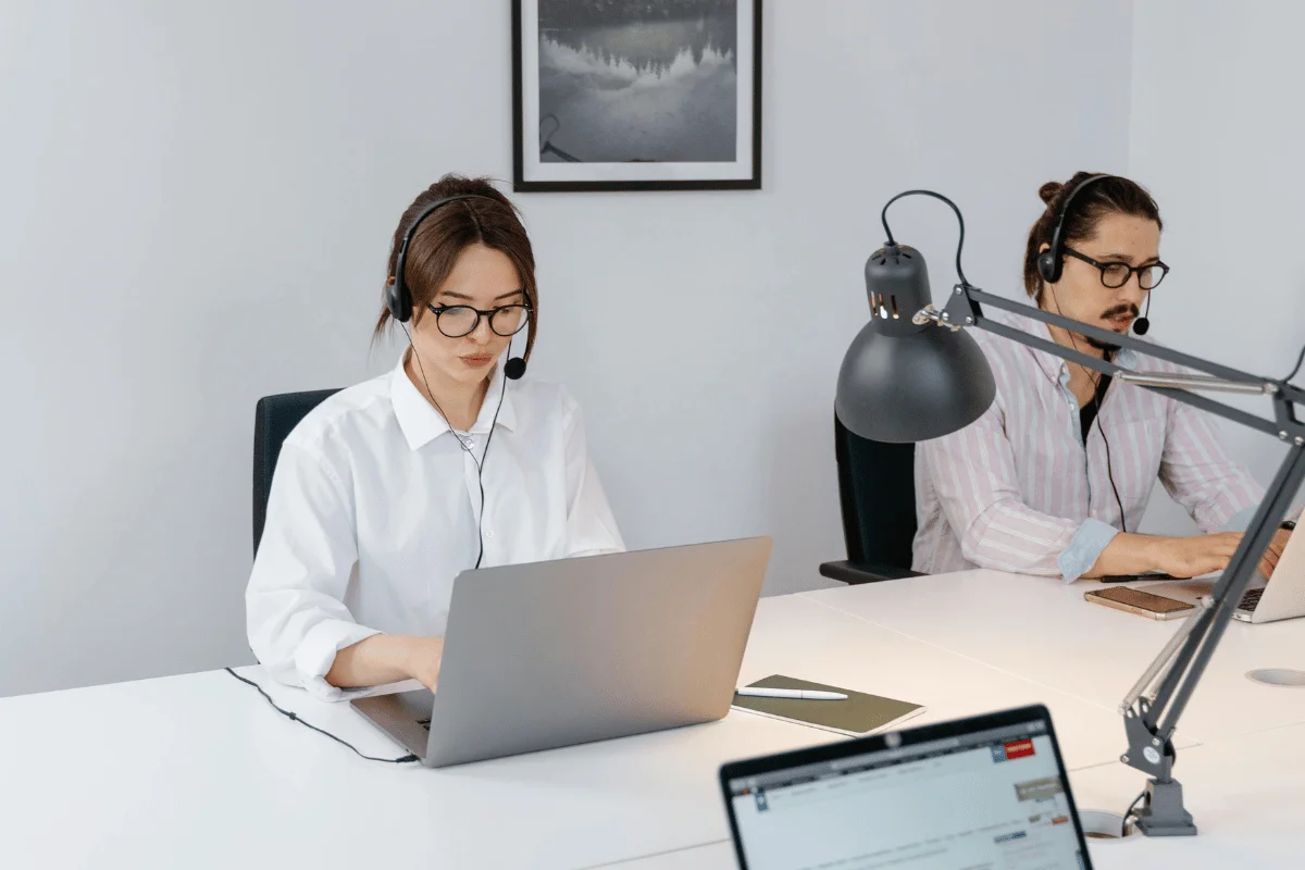 Two individuals wearing headphones work on laptops at a modern workspace with a desk lamp and a framed picture on the wall.