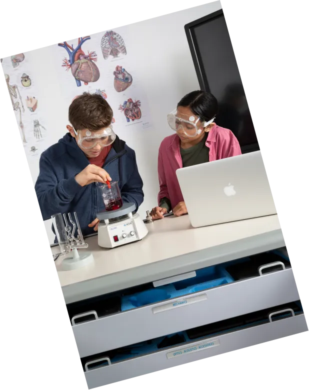 Two students in safety goggles heating a solution on a hotplate stirrer beside a laptop, with ATP Mobile Lab drawers open below