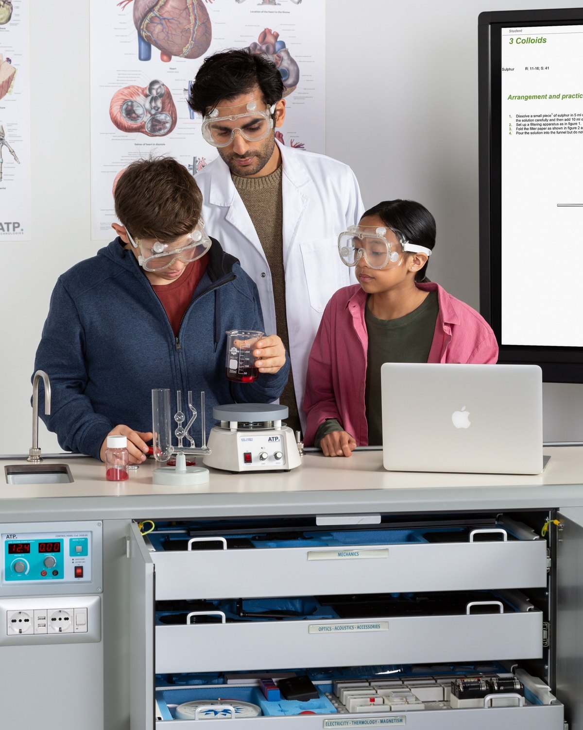 A teacher and two students in safety goggles running a chemistry experiment at the ATP Mobile Lab, with the experiment instructions on the screen behind