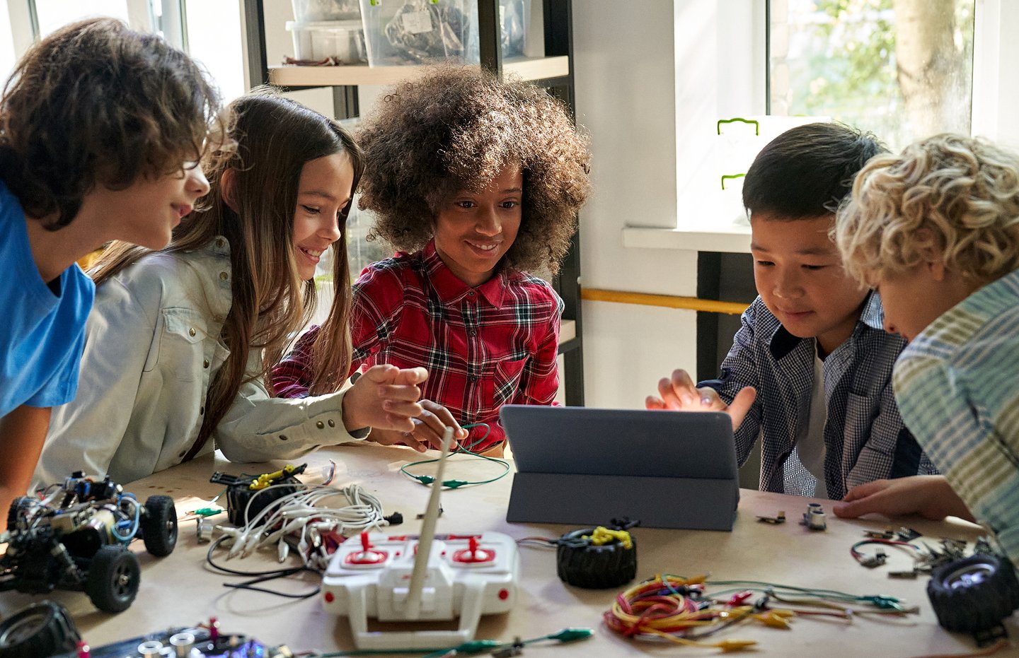 Five schoolchildren gathered around a tablet, building and testing robotics kits on a classroom table