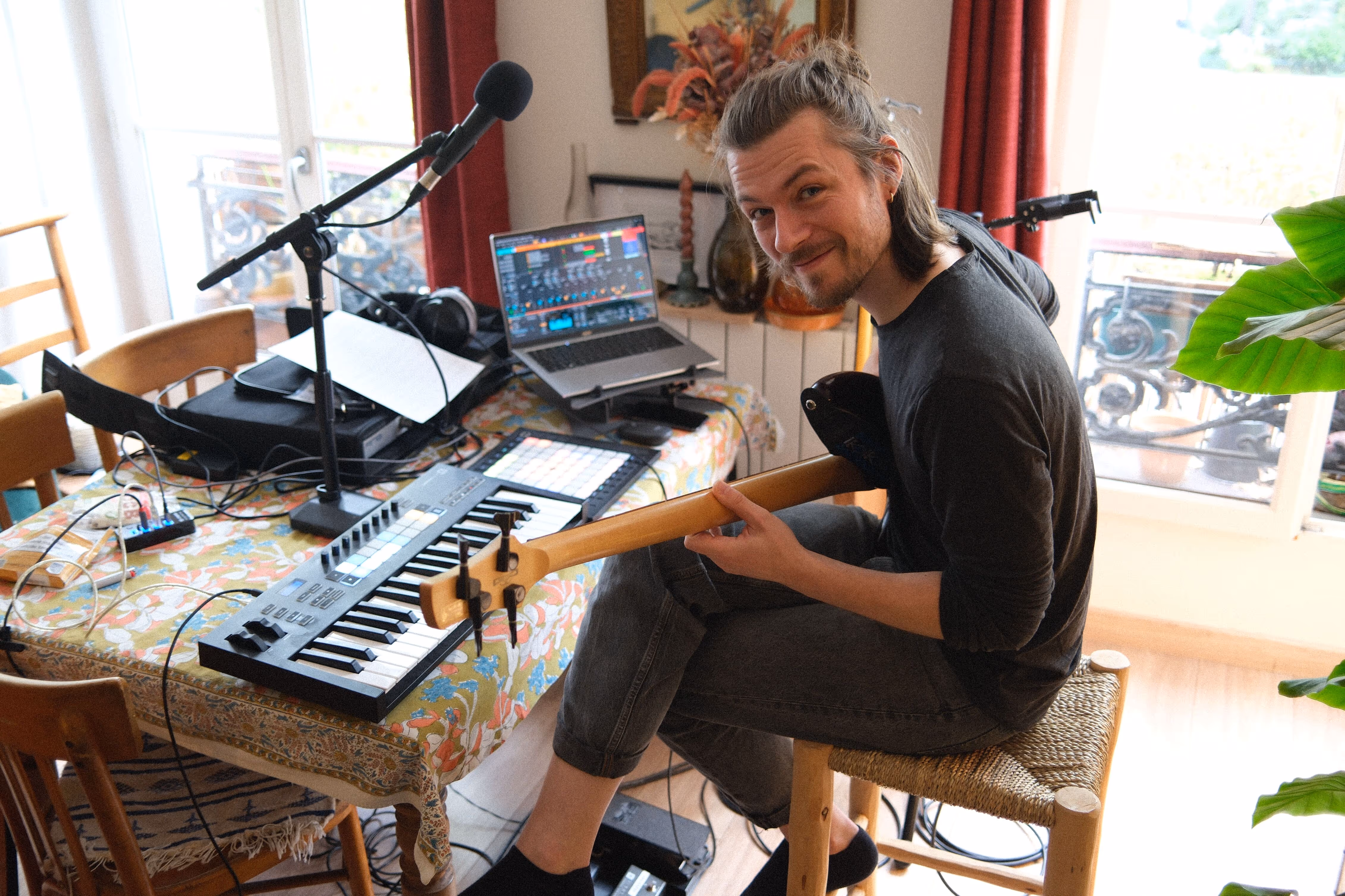 Un homme souriant tient une basse, assis devant une table couverte d'équipements audio et d'un clavier MIDI dans un salon
