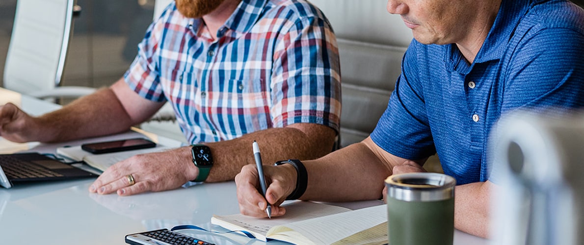 Two men sitting at a table, one typing on a laptop and the other writing in a notebook.
