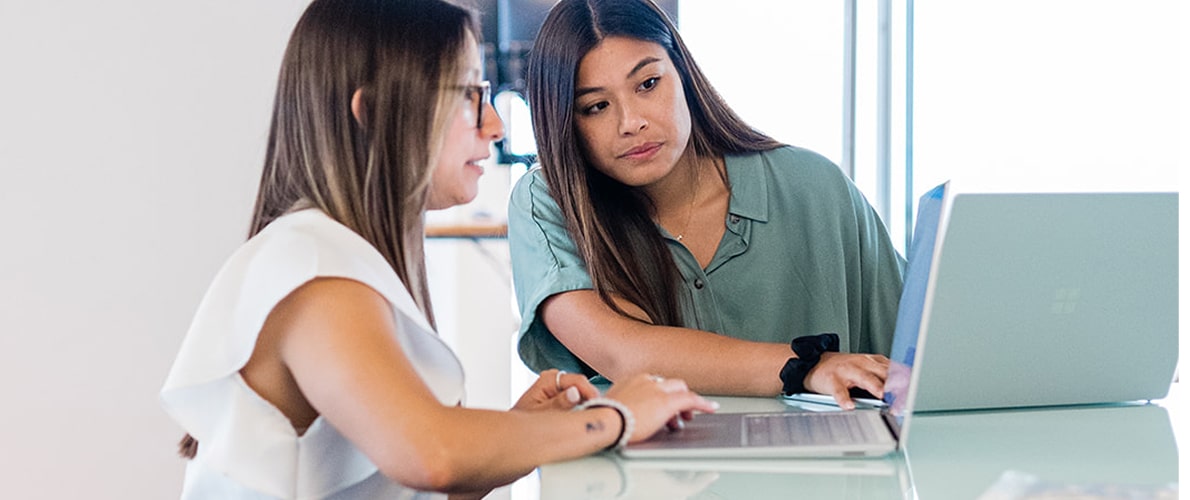 Two women working together and looking at laptops on a glass table in a bright office.