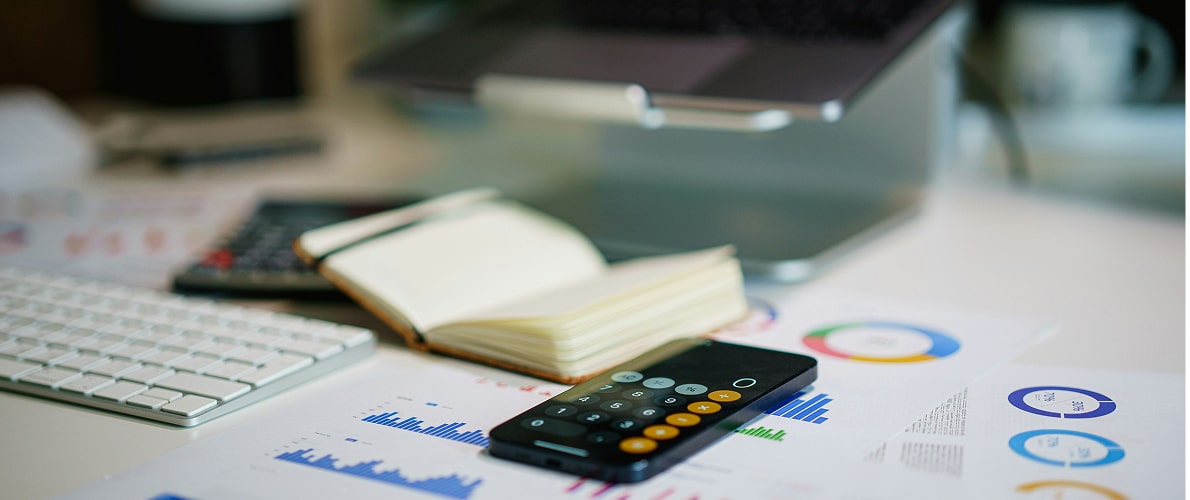 Smartphone calculator app, open notebook, keyboard, and financial charts on a desk with a laptop in the background.