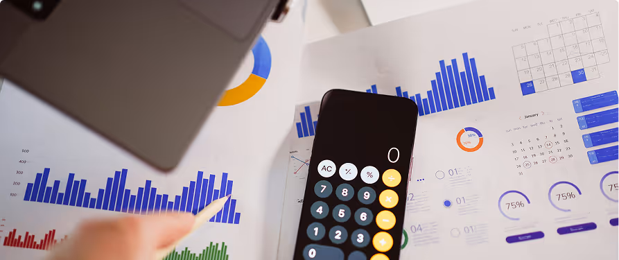Calculator on papers displaying various colorful business charts and graphs with a person's hand holding a pencil.