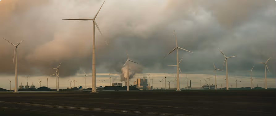 Wind turbines in the foreground with a factory emitting smoke in the background under a cloudy sky.