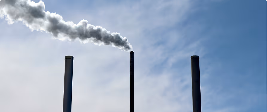 Three tall industrial smokestacks against a blue sky, with the center smokestack releasing thick white smoke.