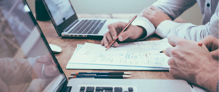 Two people collaborating over a document with notes, surrounded by open laptops on a wooden table.