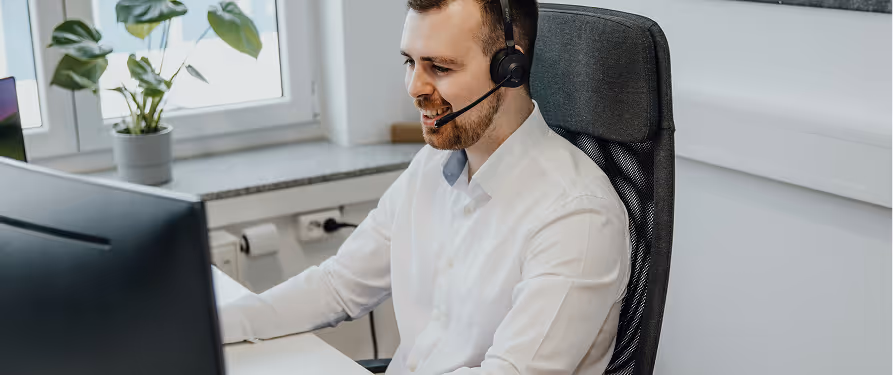 Smiling man in white shirt wearing headset and sitting at a desk with a computer monitor in a bright office.