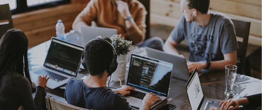 Group of young professionals collaborating with laptops around a wooden table in a casual workspace.