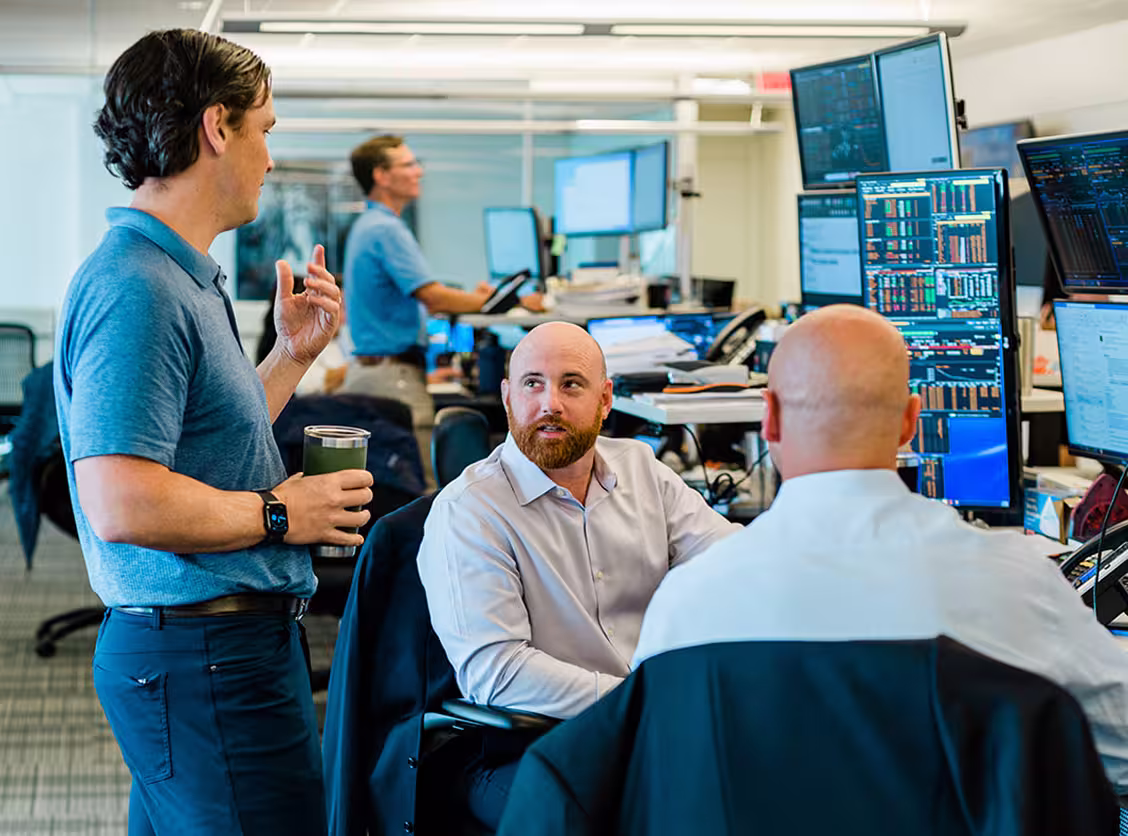 Three men working in an office with multiple computer monitors displaying data and charts.