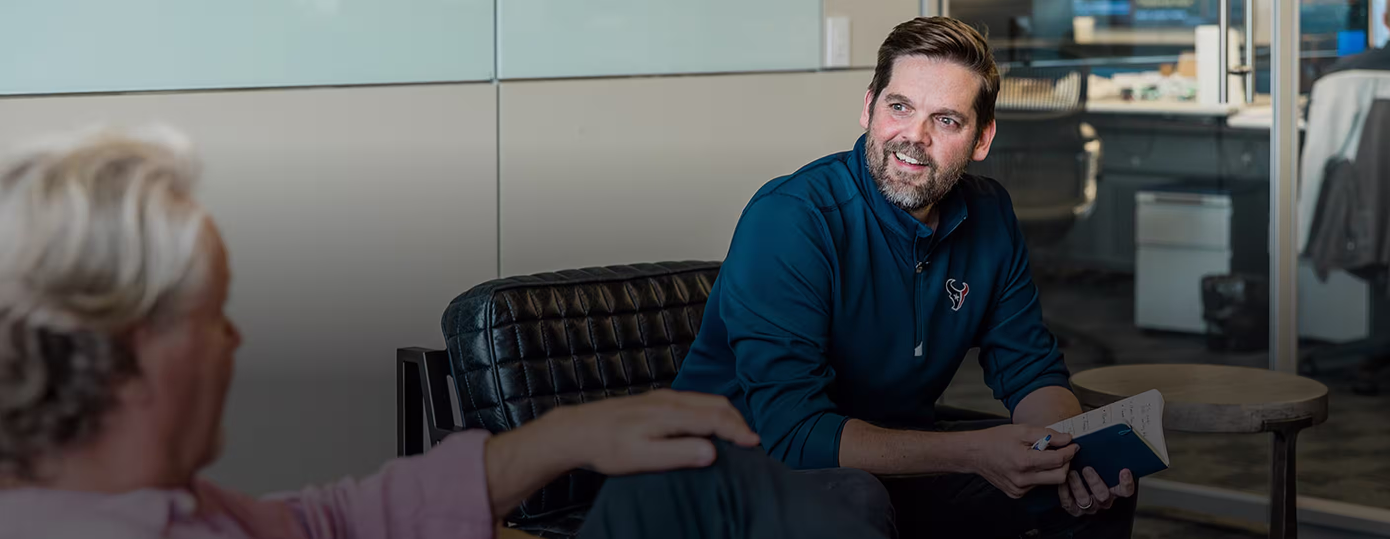 Man in blue pullover smiling and holding a notebook while talking to another person in an office.